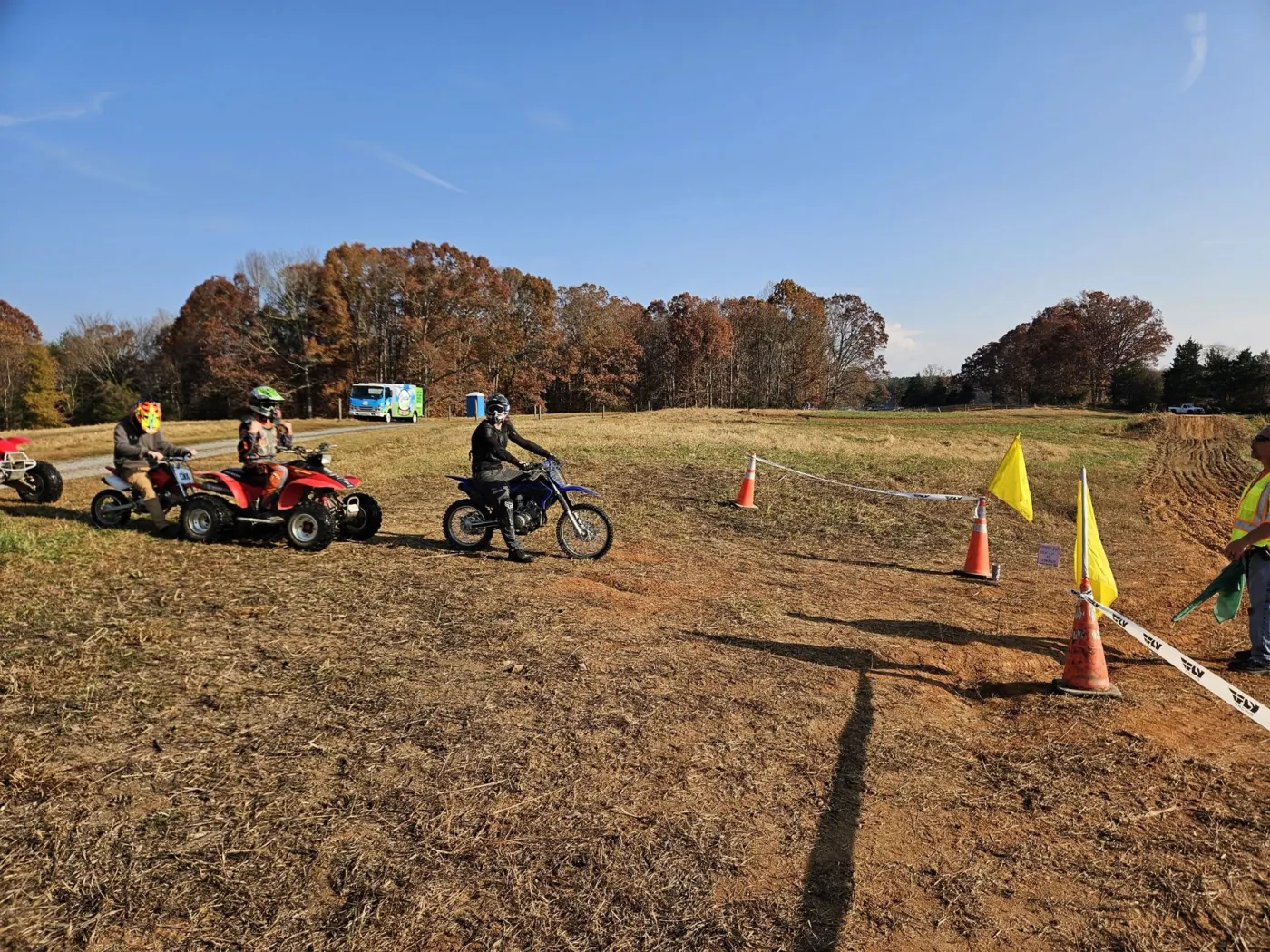 Youth riders on ATVs and a dirt bike lined up at the track entrance with flaggers